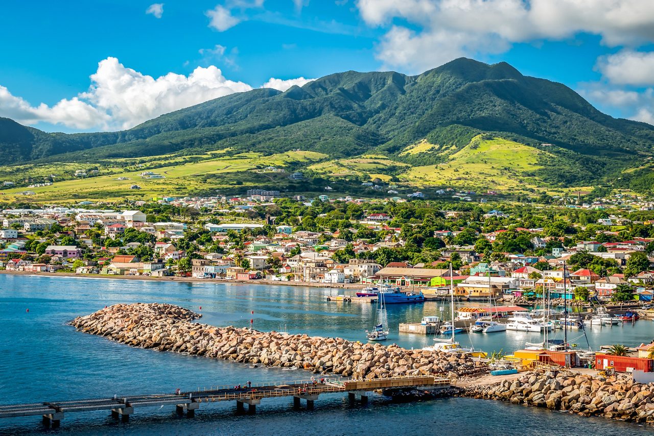 Coastal town of Basseterre in St Kitts with colorful buildings along the shore, a marina full of boats and a rock breakwater in the foreground, backed by lush green mountains under a blue sky.