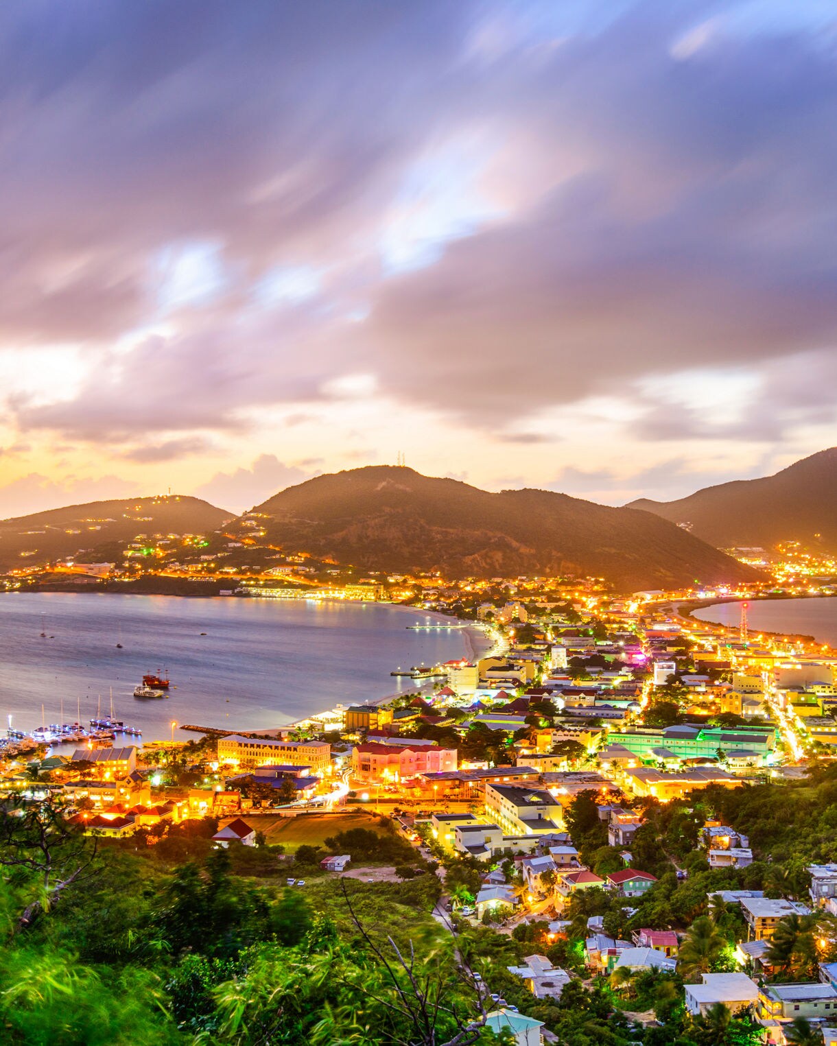 A panoramic sunset view of Philipsburg on a small Caribbean island with bright city lights curving along two bays. Mountains rise in the background under streaked clouds while boats rest in calm water and green hills fill the foreground.