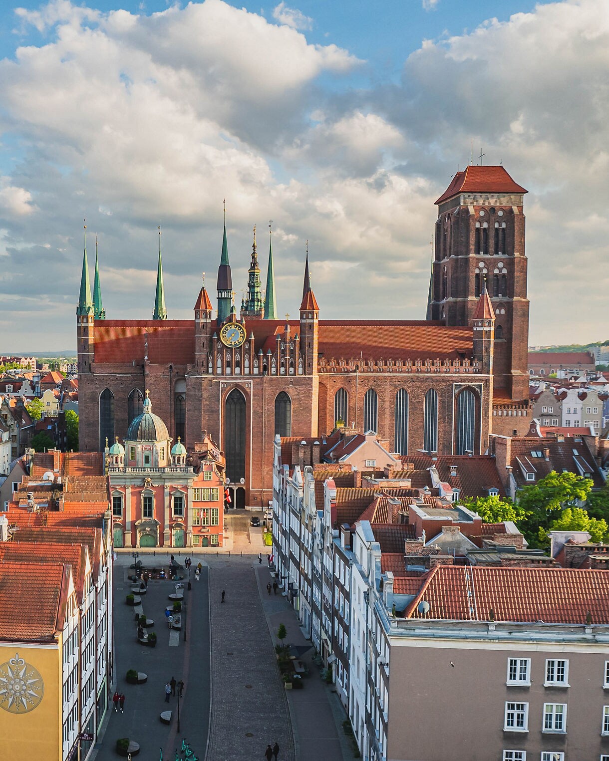Aerial view of St. Mary’s Basilica in Gdańsk, Poland, featuring its massive brick walls, tall spires and large clock, surrounded by colorful historic townhouses with red-tiled roofs.