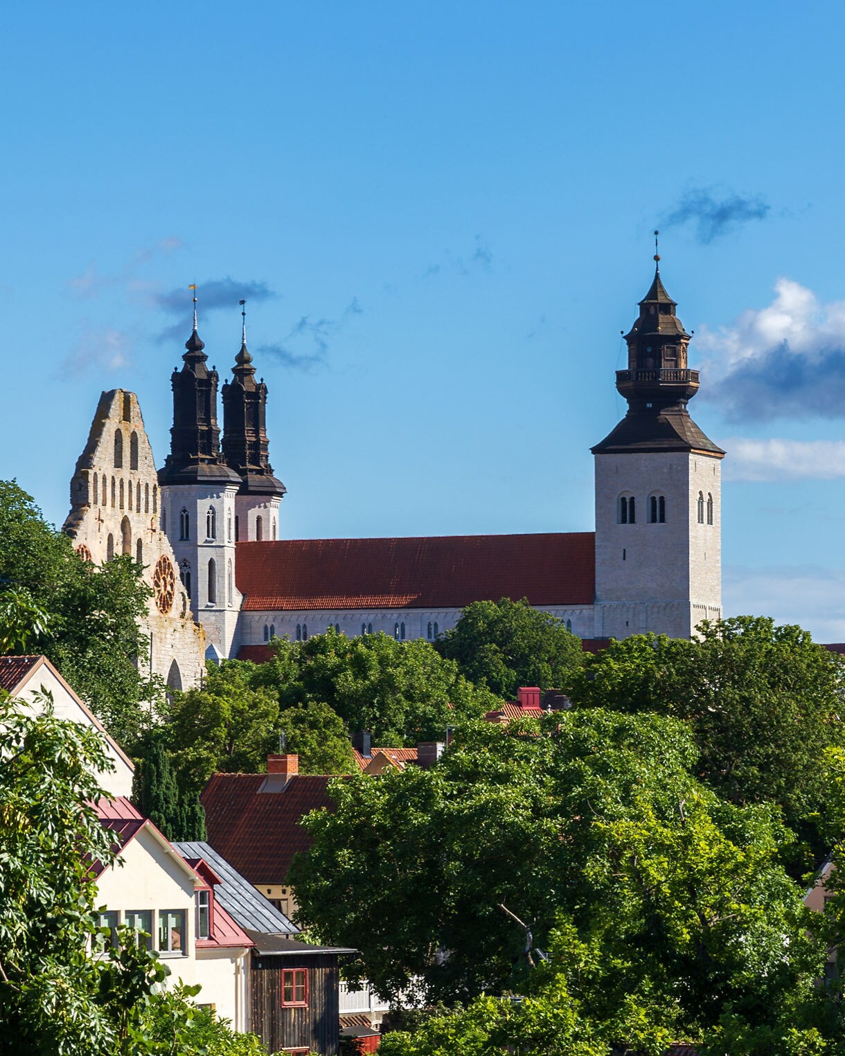 St. Mary’s Cathedral in Visby, Gotland, Sweden, with twin dark spires and a central tower rising above red rooftops and green trees under a clear blue sky.