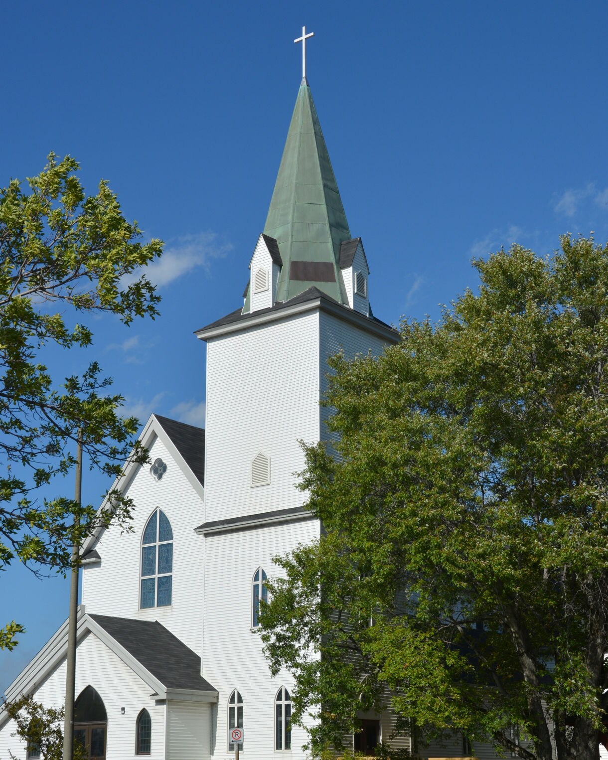 White wooden church with a tall green steeple topped by a cross, surrounded by leafy trees on a sunny day.