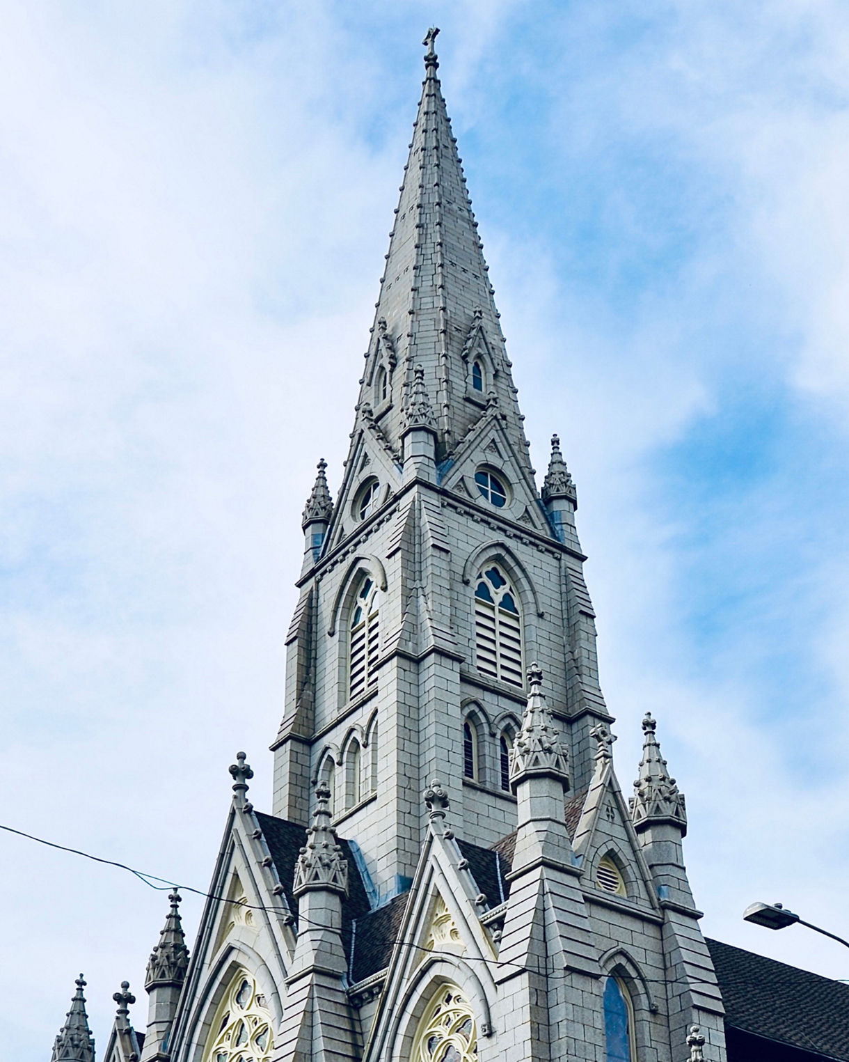 Tall Gothic-style cathedral with a sharply pointed spire, ornate stonework, arched windows and wooden doors set beneath a bright blue sky.