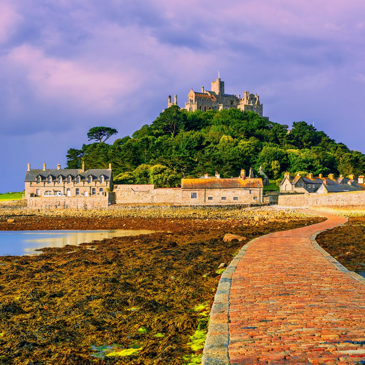Stone pathway leading across tidal flats to St Michael’s Mount in Cornwall, with a medieval castle perched on a green hill under a colorful sky.