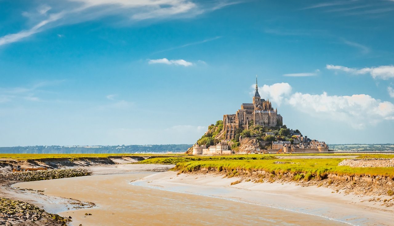 View of Mont Saint-Michel, a medieval abbey and village perched on a rocky island, surrounded by tidal flats under a bright blue sky.