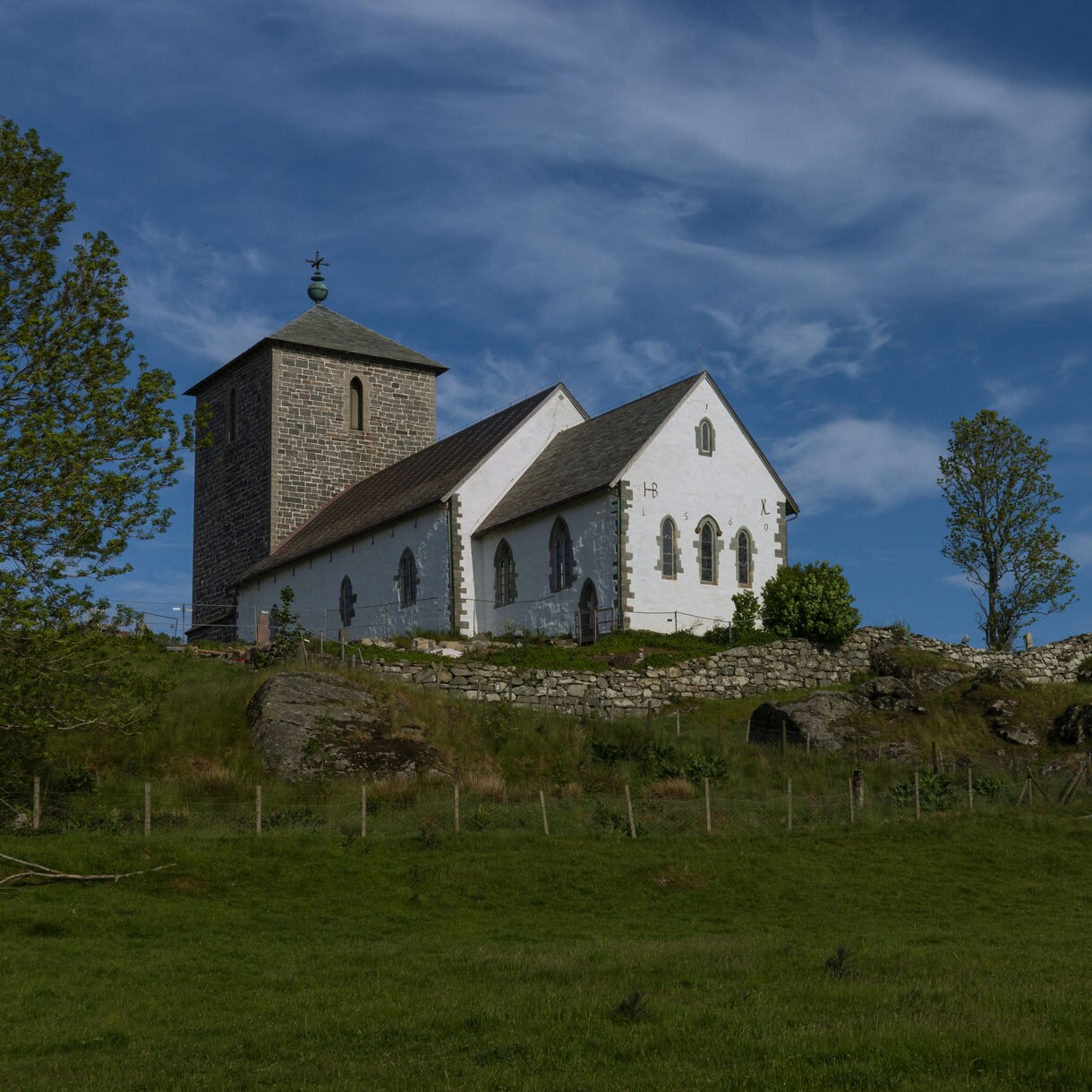 St. Olav’s Church, a white stone medieval church with a square tower, sits on a grassy hillside surrounded by trees and stone walls under a bright blue sky.