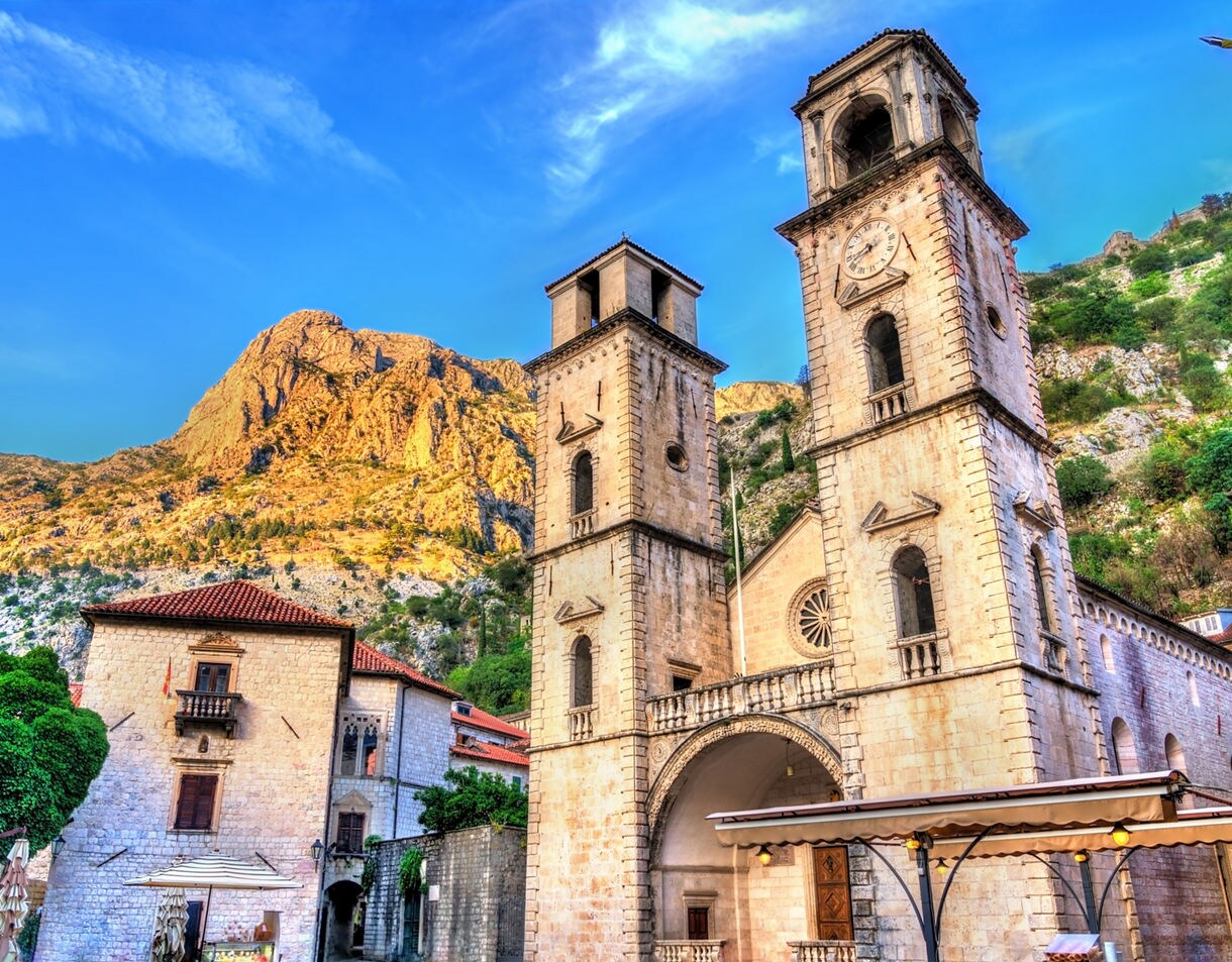St. Tryphon Cathedral in Kotor, Montenegro, with twin stone bell towers and a clock face, framed by old town buildings and steep rocky mountains in the background.