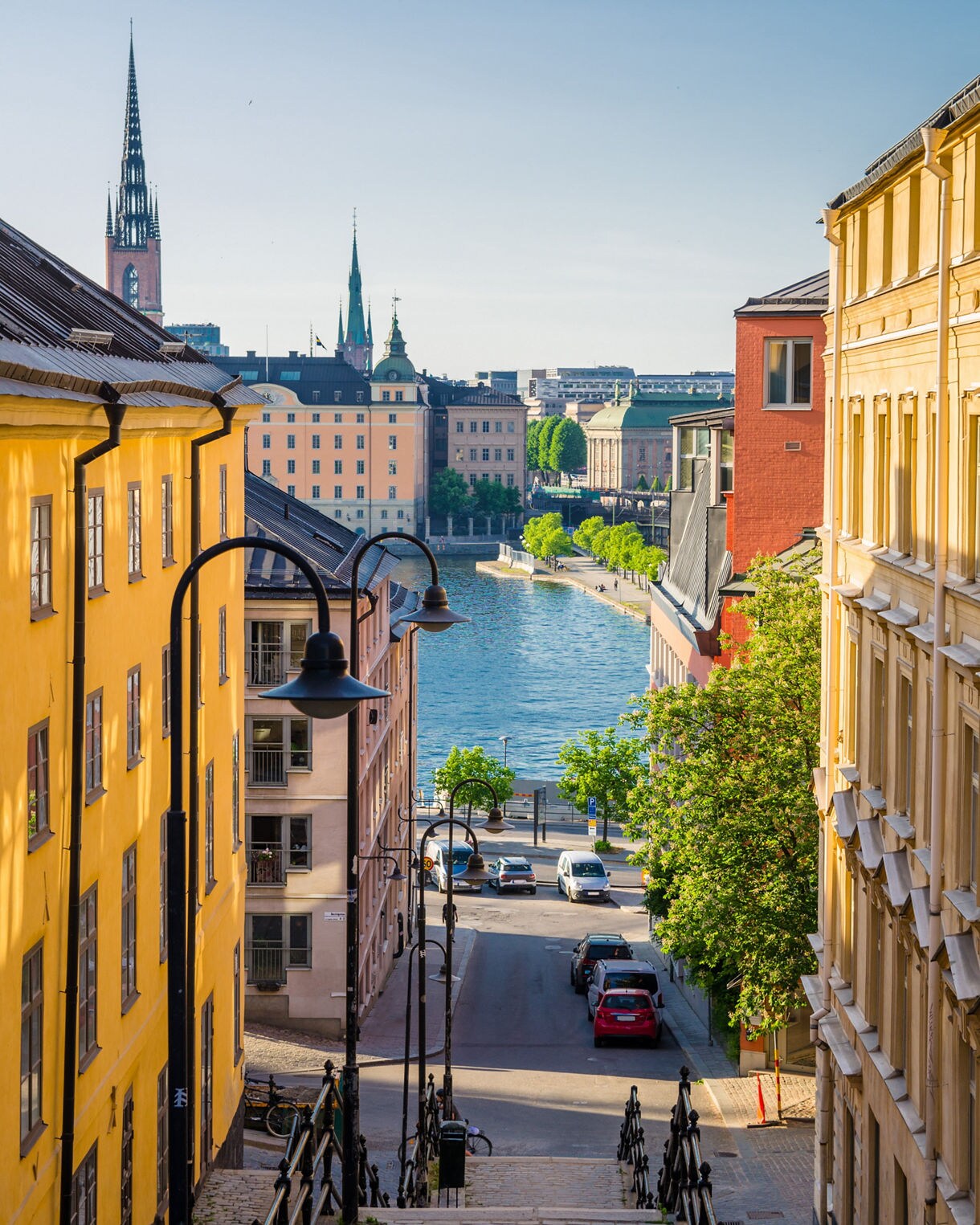 View from a cobblestone stairway lined with colorful yellow and red buildings, leading down to Lake Mälaren with church spires and historic Stockholm architecture in the background.