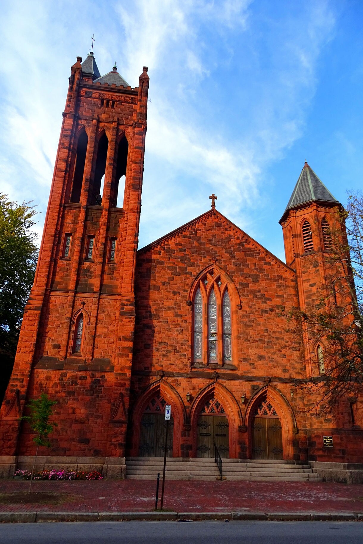 A tall red-stone Gothic church lit by warm evening light, featuring two towers, pointed arches and a trio of wooden doors framed by ornate stonework beneath a bright blue sky.