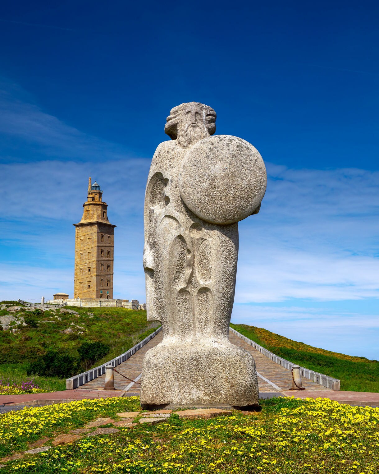 Stone statue of Breogán holding a round shield with the Tower of Hercules in the background on a grassy coastal hill under a bright blue sky.
