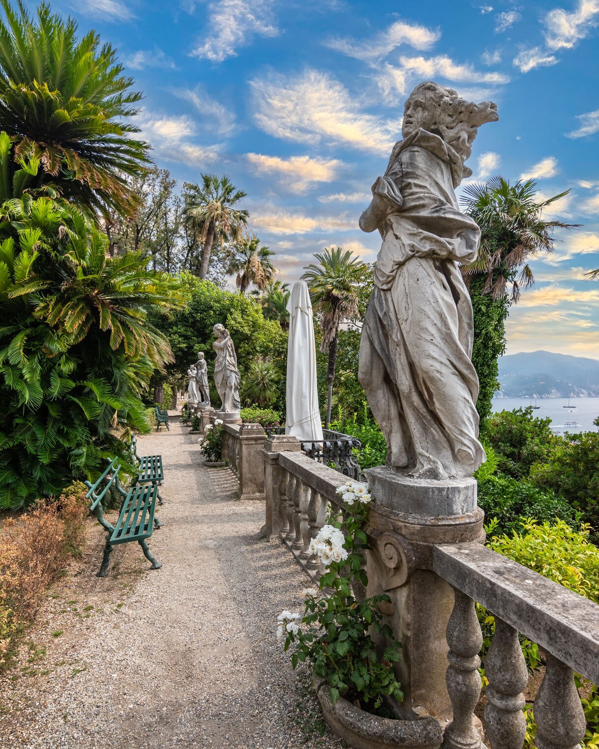 A pathway in a lush garden overlooking the sea, lined with classical stone statues and green benches, surrounded by palm trees and flowering plants.