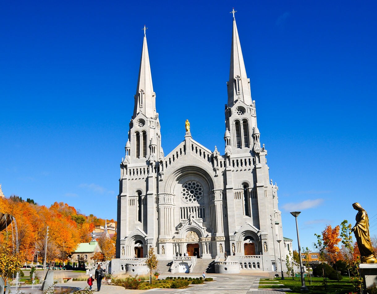 Grand stone basilica with two tall spires at Sainte-Anne-de-Beaupré, framed by fall foliage, statues and a fountain under a bright blue sky.