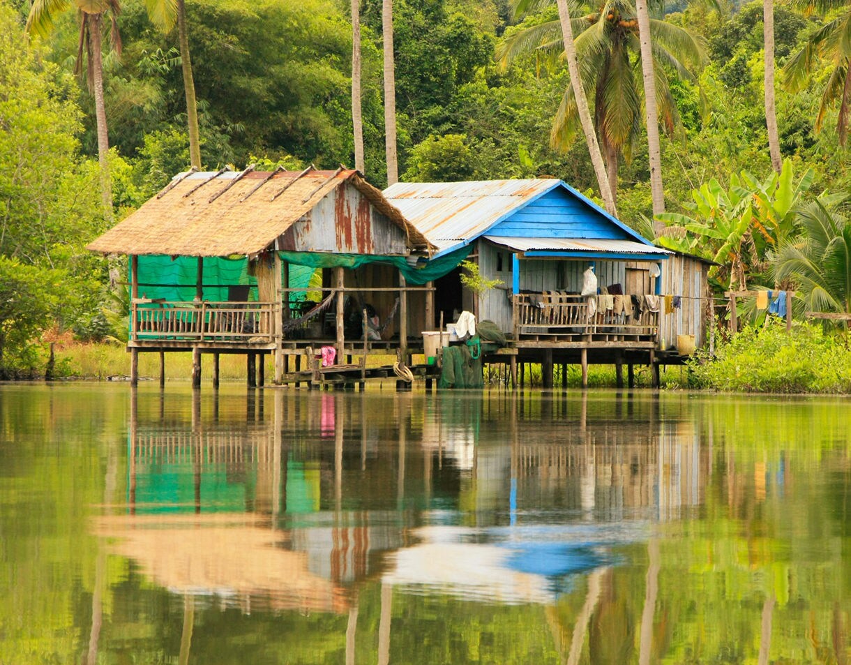 Small wooden stilt houses with tin and thatched roofs stand over still water surrounded by dense tropical trees and palms.
