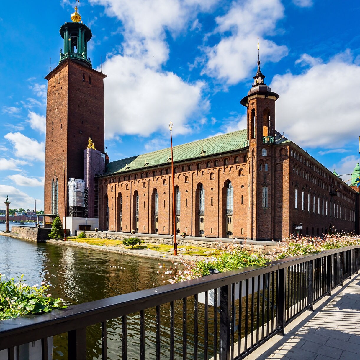 Stockholm City Hall with its tall brick tower topped by golden crowns, set beside the waterfront on a bright sunny day with flowers in the foreground.