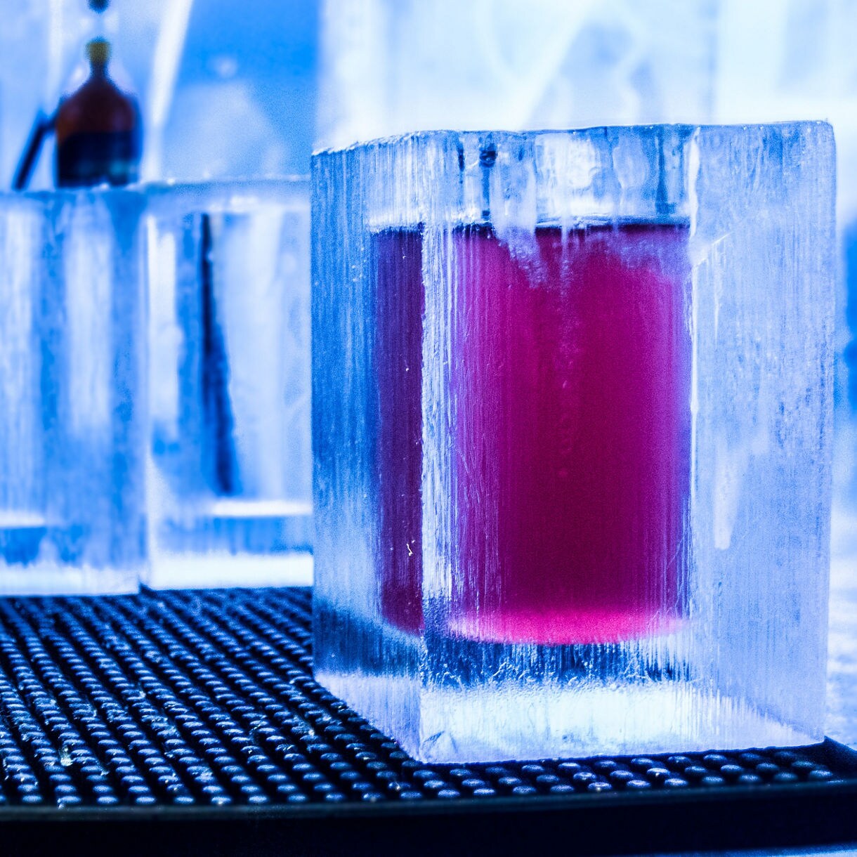 Close-up of a red cocktail served in a square glass made of ice at Stockholm’s Ice Bar, with bottles blurred in the frosty blue background.