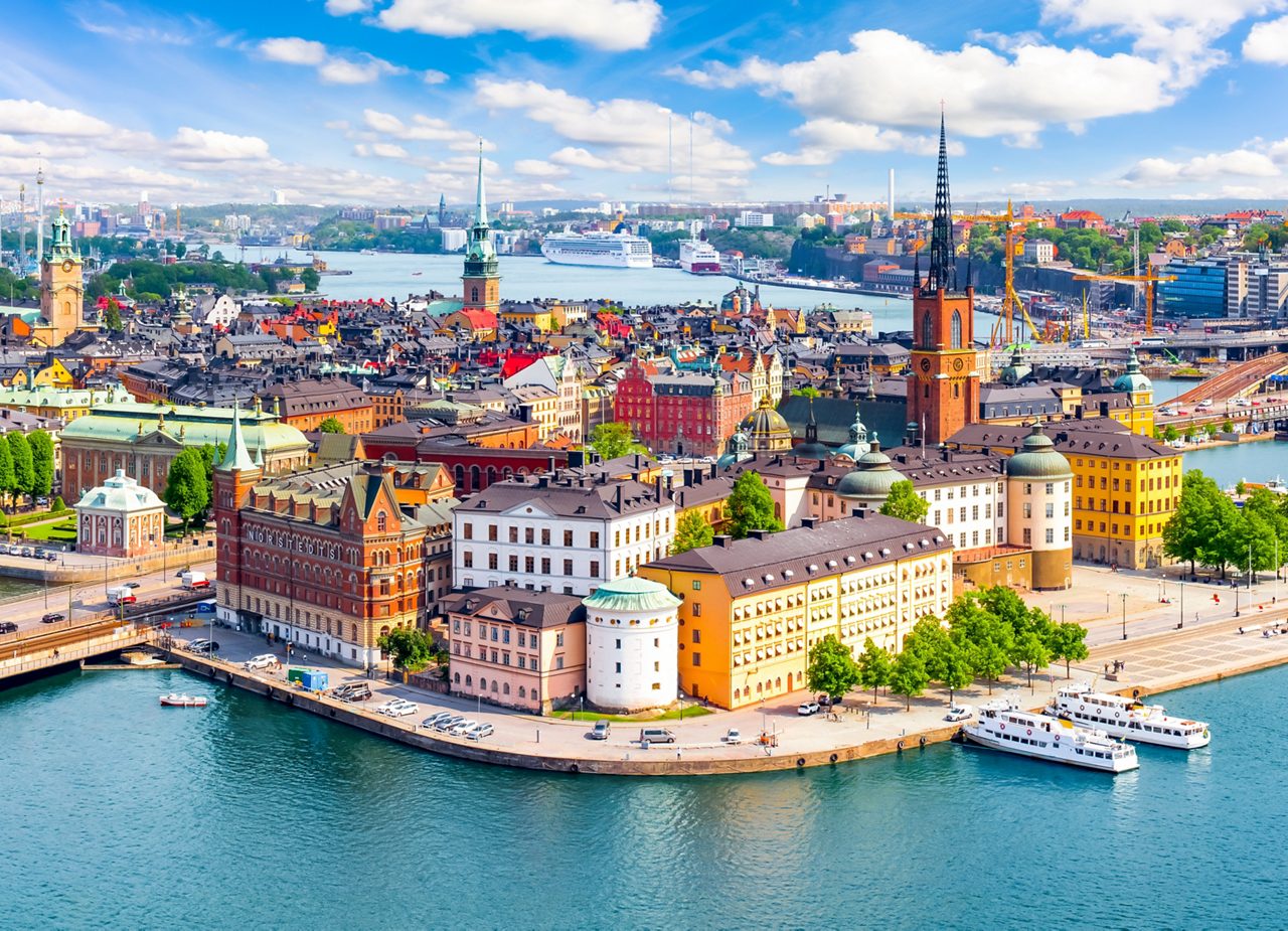 Aerial view of Stockholm’s historic Gamla Stan with colorful buildings, church spires and boats docked along the waterfront on a sunny day.