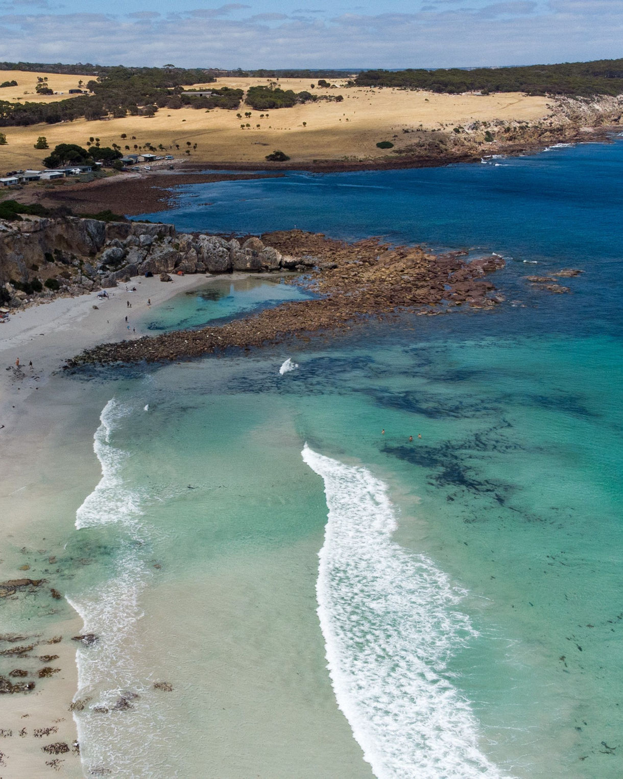 Aerial view of Stokes Bay with clear turquoise water, white sand, rocky shoreline and rolling golden hills beyond.