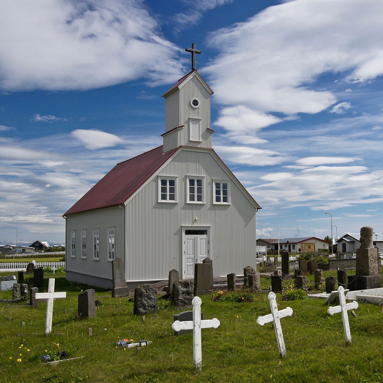 Small white wooden church with a red roof and bell tower, surrounded by a grassy cemetery with stone and wooden crosses under a partly cloudy sky in Stokkseyri, Iceland.