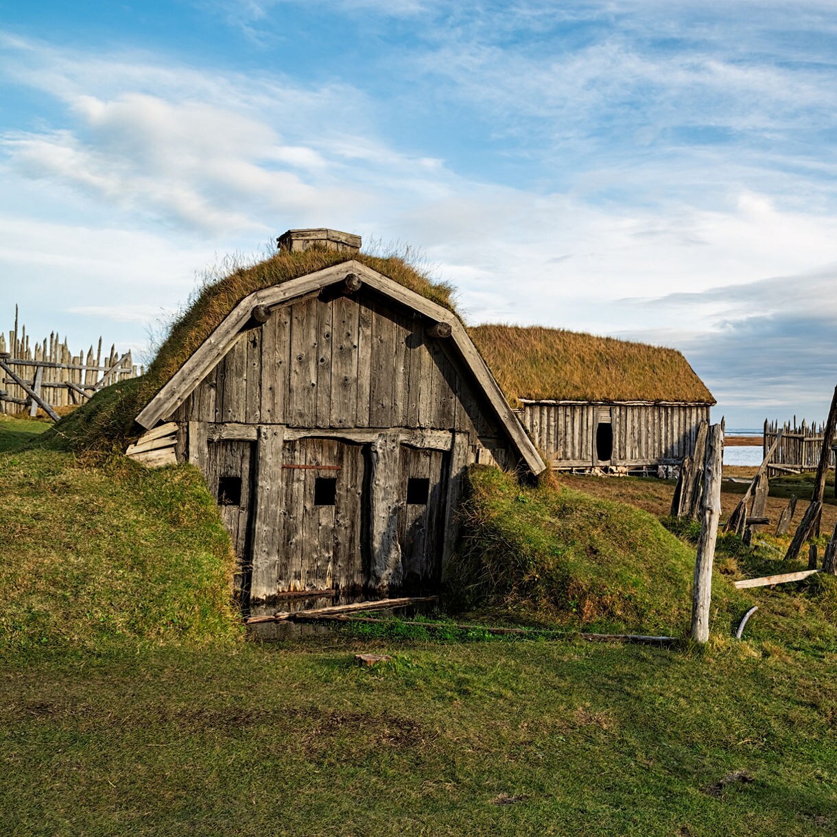 Old wooden Viking-style buildings with grass-covered roofs set on a grassy coastal plain under a partly cloudy sky.