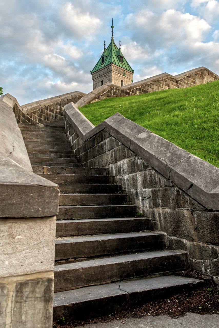 Old stone steps rising between fortress walls toward a small tower with a green roof on a grassy hill under a cloudy sky.