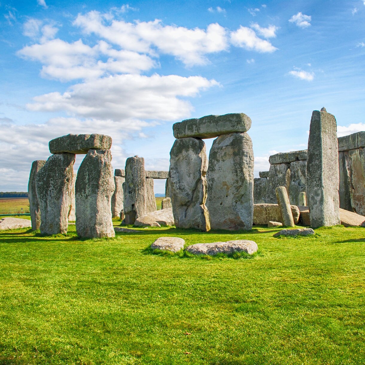 Stonehenge stone circle in Wiltshire, featuring massive upright sarsen stones topped with horizontal lintels, set against green grass and a partly cloudy blue sky.