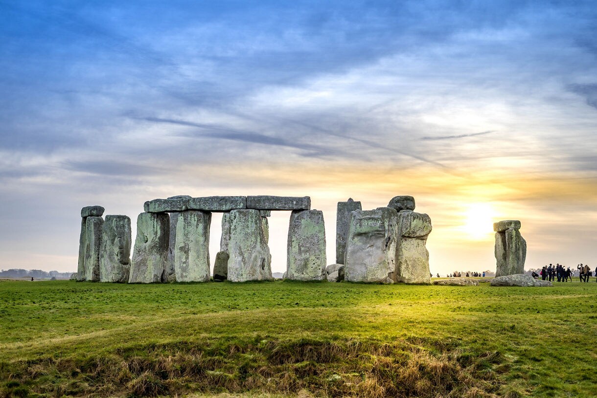 Stonehenge at dusk with massive upright stones topped by lintels, glowing in golden light as the sun sets behind a partly cloudy sky.
