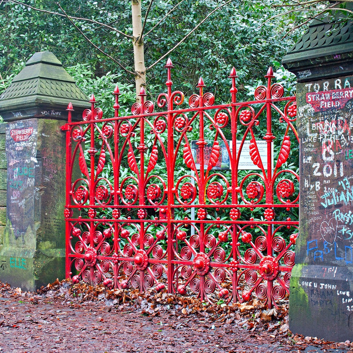 Bright red ornate gates at Strawberry Field in Liverpool, with graffiti-covered stone pillars and trees in the background.