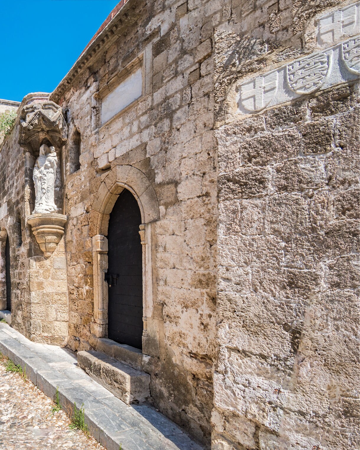 Narrow cobblestone street lined with medieval stone buildings and arched passageways under a clear blue sky in the Old Town of Rhodes, Greece.