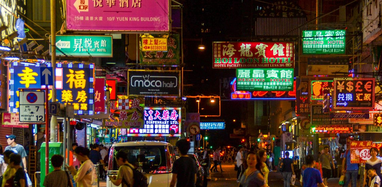 Crowded Hong Kong street at night illuminated by colorful neon signs in English and Chinese above pedestrians and cars.