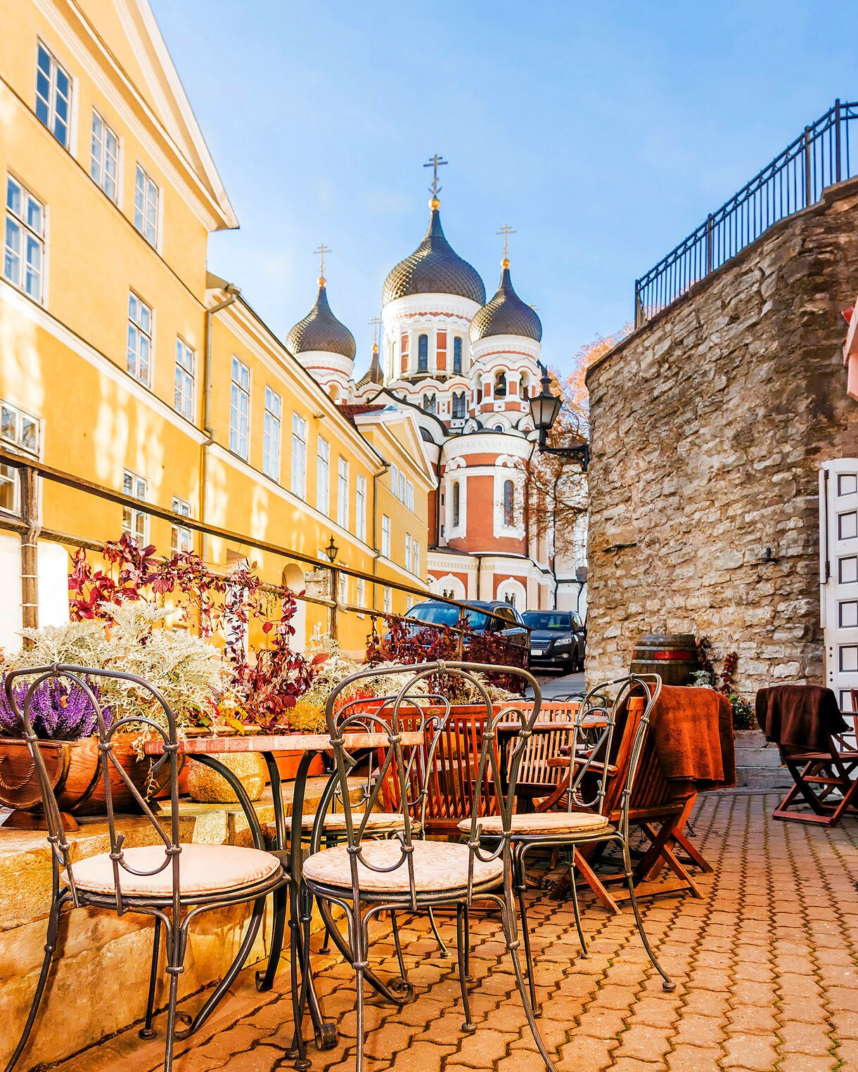 Outdoor café in a cobbled street of Tallinn, Estonia, with wrought iron chairs, yellow buildings and the onion domes of Alexander Nevsky Cathedral rising in the background.