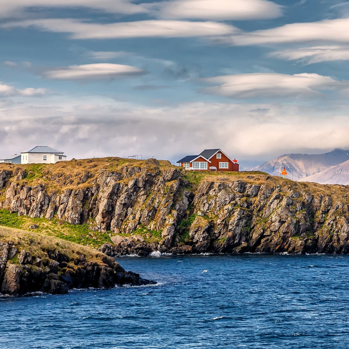 A cluster of small houses, including a red one, sitting atop rocky coastal cliffs with deep blue water below and misty mountains rising in the background beneath layered clouds.