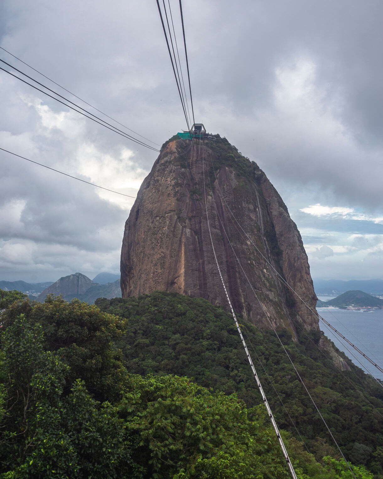 View of Sugarloaf Mountain with cable car lines leading to its summit, framed by lush green forest and an overcast sky.