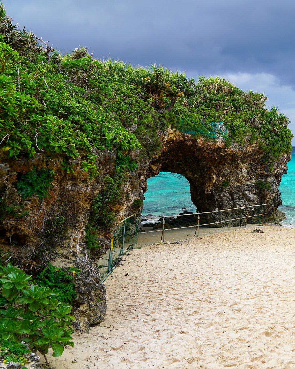 Sandy shore at Sunayama Beach featuring a lush, vegetation-covered rock arch on the left and clear turquoise water under a moody, overcast sky.