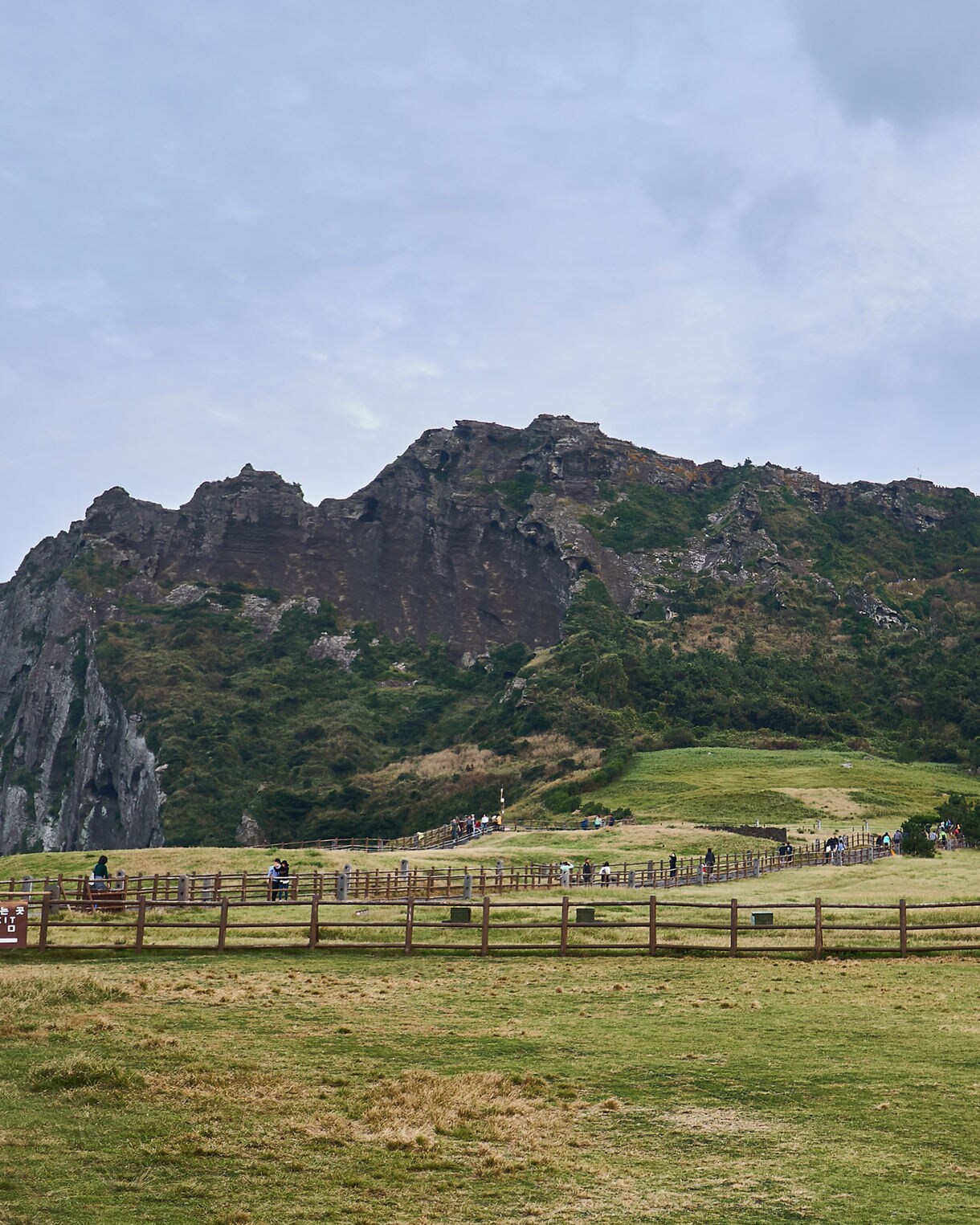 Grassy path leading toward the rocky slopes of Seongsan Ilchulbong on Jeju Island, South Korea, under a cloudy blue sky.