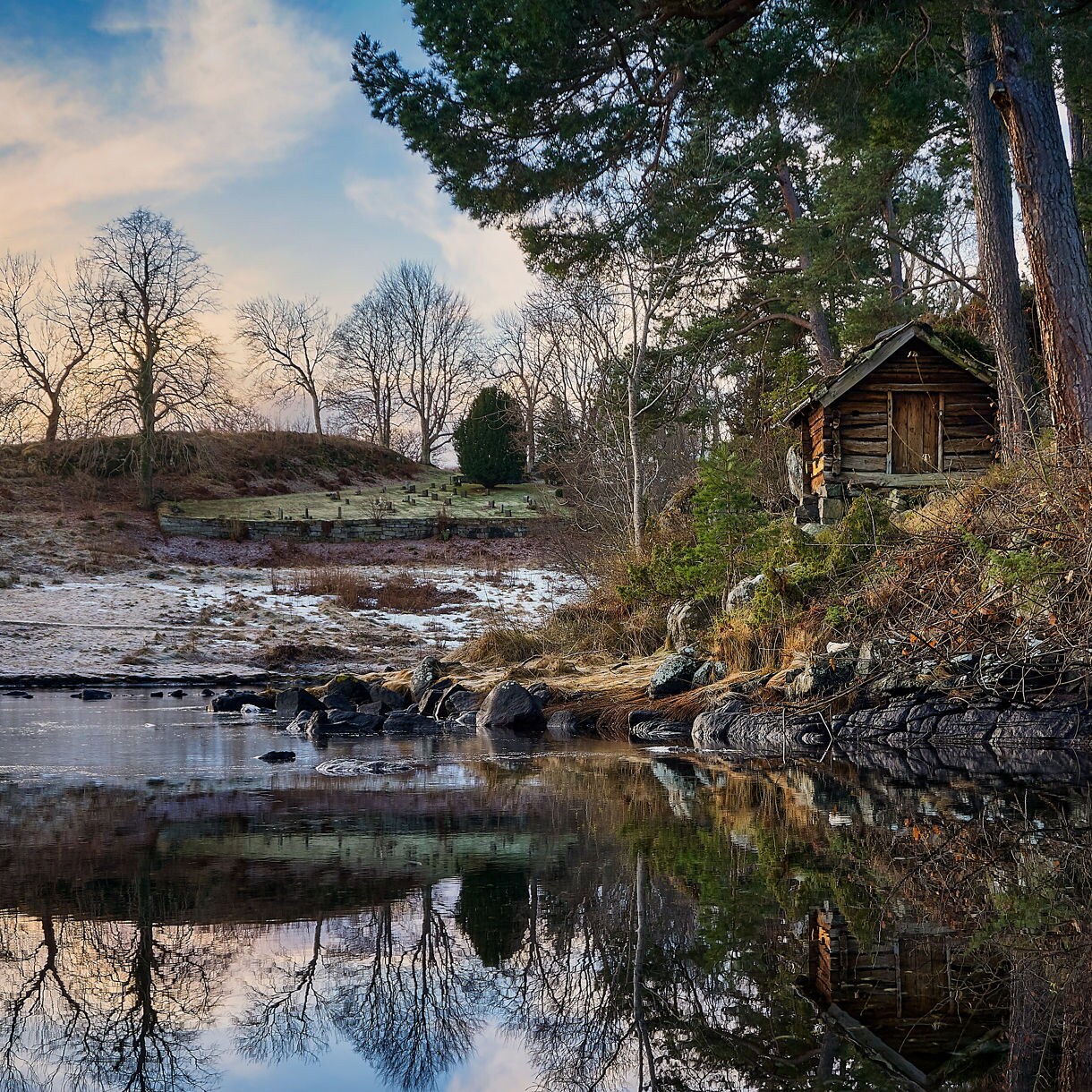 Traditional wooden cabin on a forested riverbank at Sunnmøre Museum, Norway, reflected in the calm water with bare winter trees in the distance.