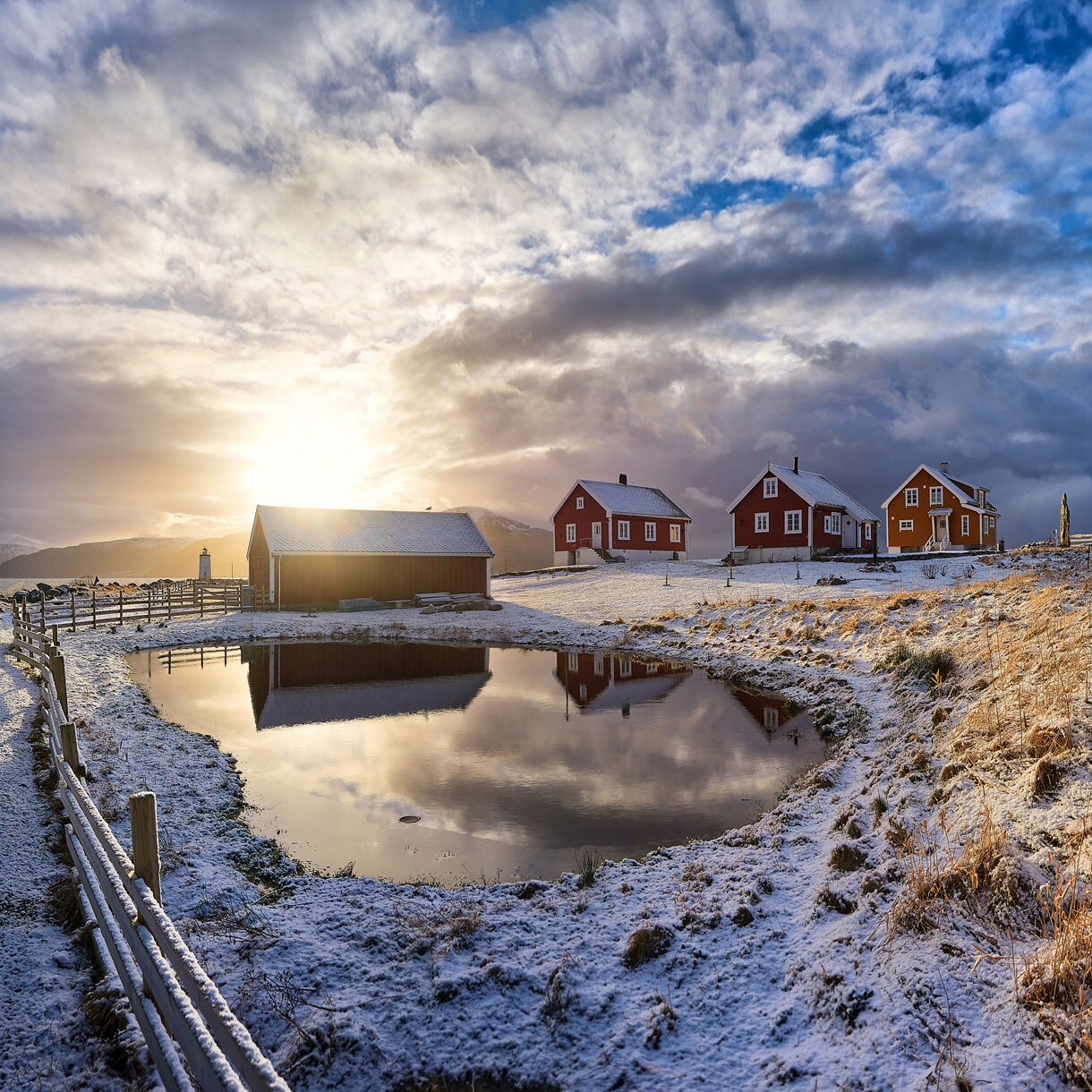 Snow-dusted landscape with red wooden houses, a barn and a reflective pond beneath dramatic winter skies on Godøy, Norway.