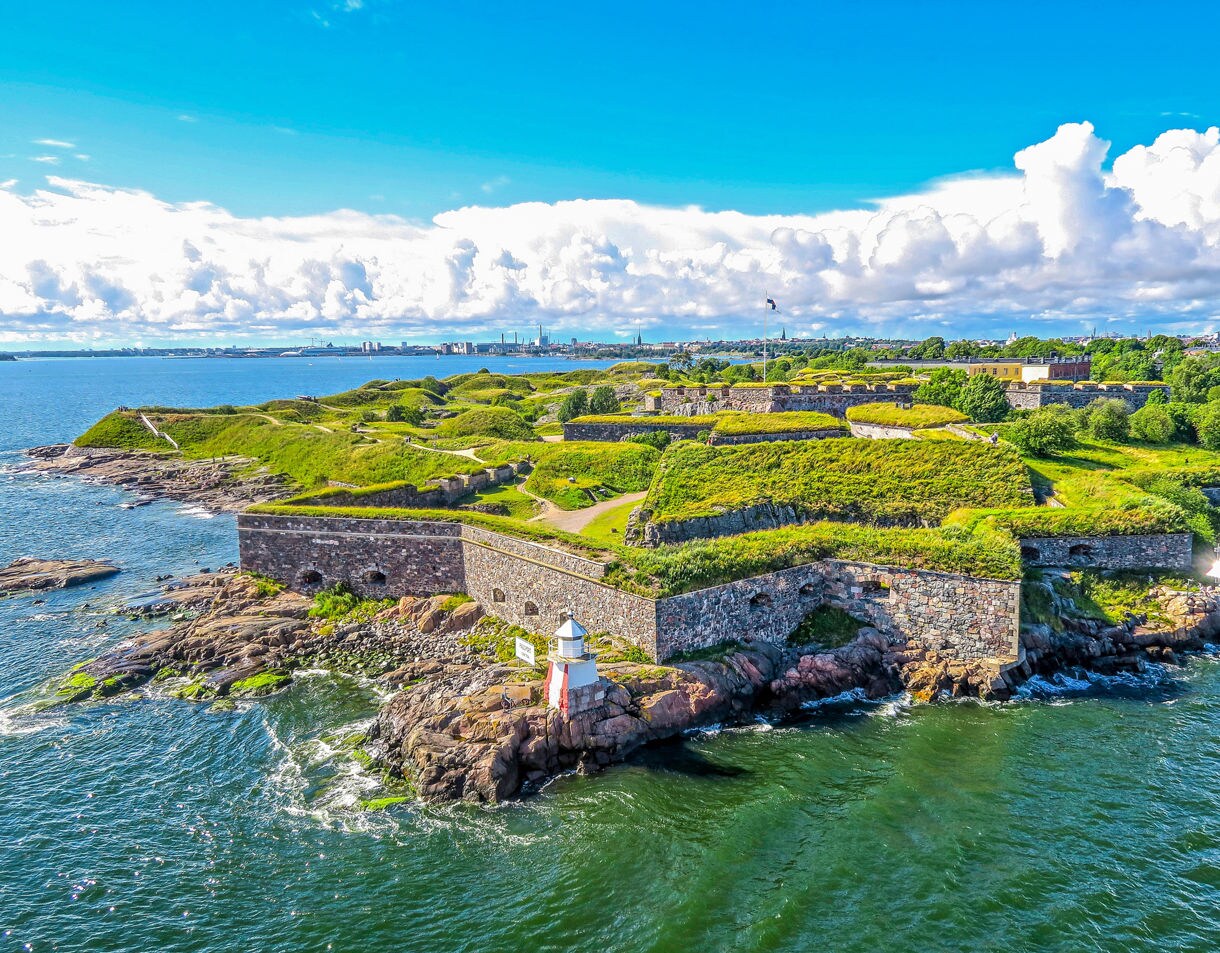 Aerial view of Suomenlinna Fortress near Helsinki, with stone walls, green grassy mounds and a small white lighthouse overlooking the blue sea.