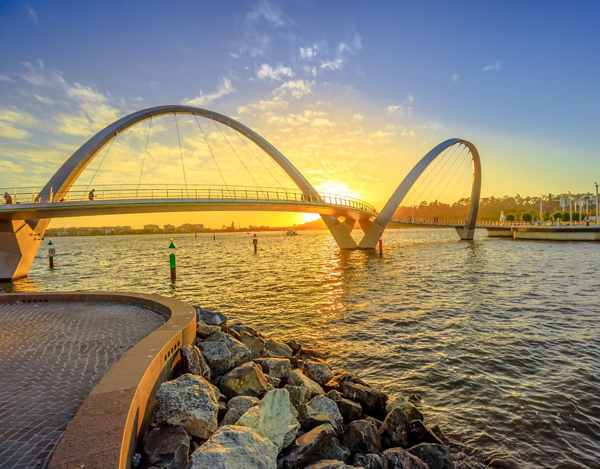 Modern double-arched pedestrian bridge spanning the Swan River at sunset with golden reflections on the water.