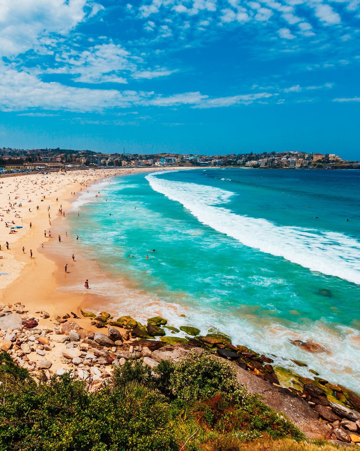 A wide view of Bondi Beach with turquoise waves rolling onto a long stretch of sandy shore filled with sunbathers, swimmers and coastal buildings under a partly cloudy blue sky.