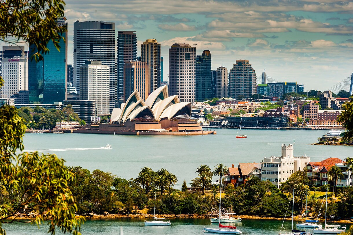 View of Sydney Harbour with the Opera House centered in front of tall skyscrapers, framed by leafy neighborhoods, palm trees and sailboats floating in the calm water under a partly cloudy sky.