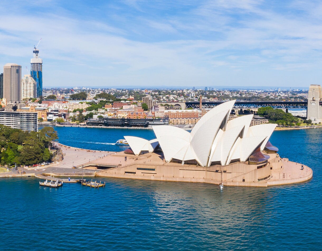 Aerial view of the Sydney Opera House with its white sail-like roofs, surrounded by blue harbor water, nearby parks and the city skyline stretching across the background.