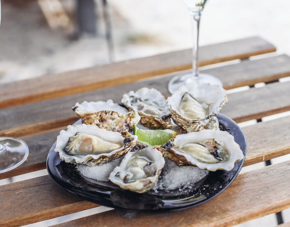 Close-up of a black plate filled with shucked oysters resting on crushed ice with lime wedges, placed on a wooden outdoor table with two glasses of white wine nearby.