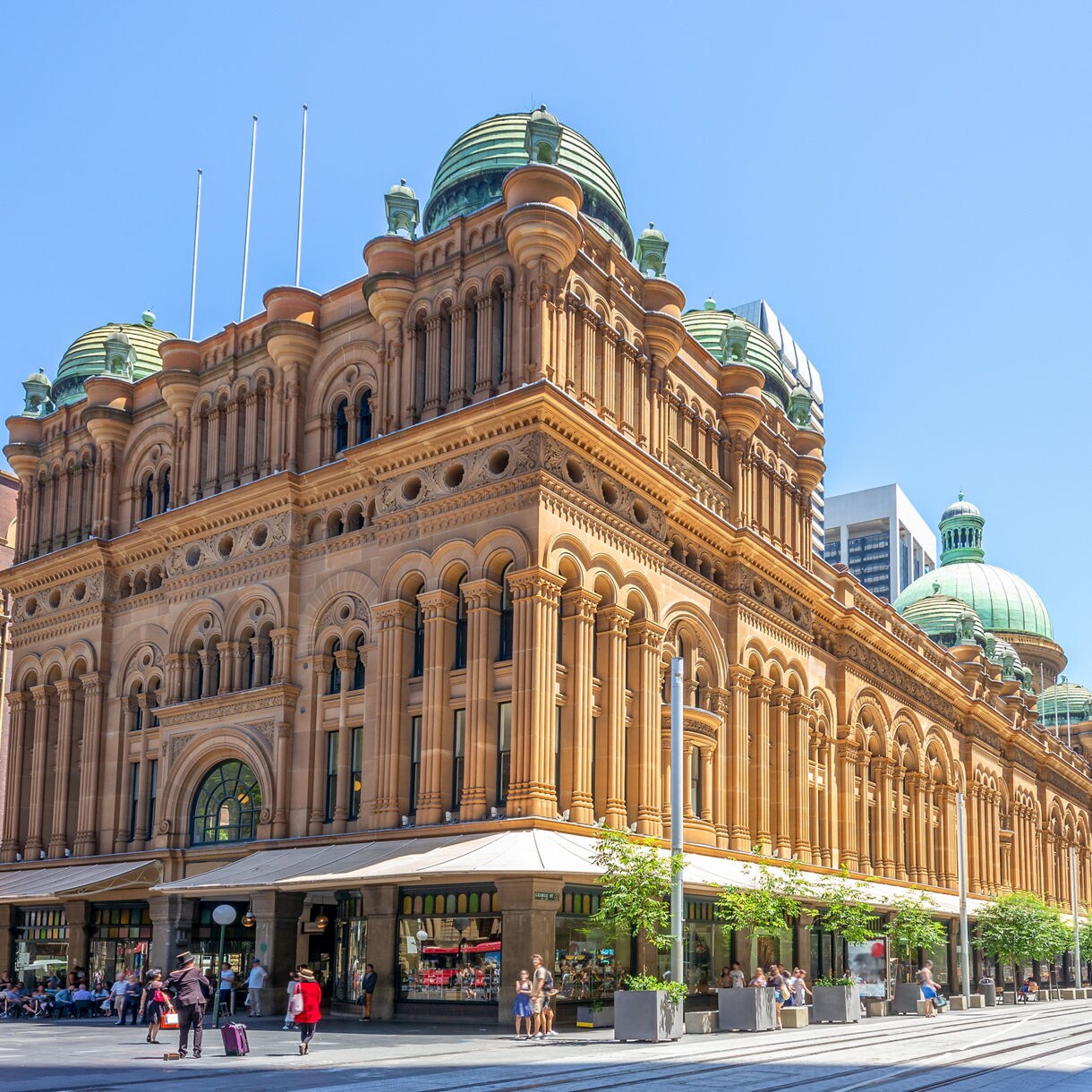 Street-level view of the Queen Victoria Building in Sydney featuring ornate sandstone arches, green domes and a busy pedestrian street with shoppers and café tables under a clear blue sky.