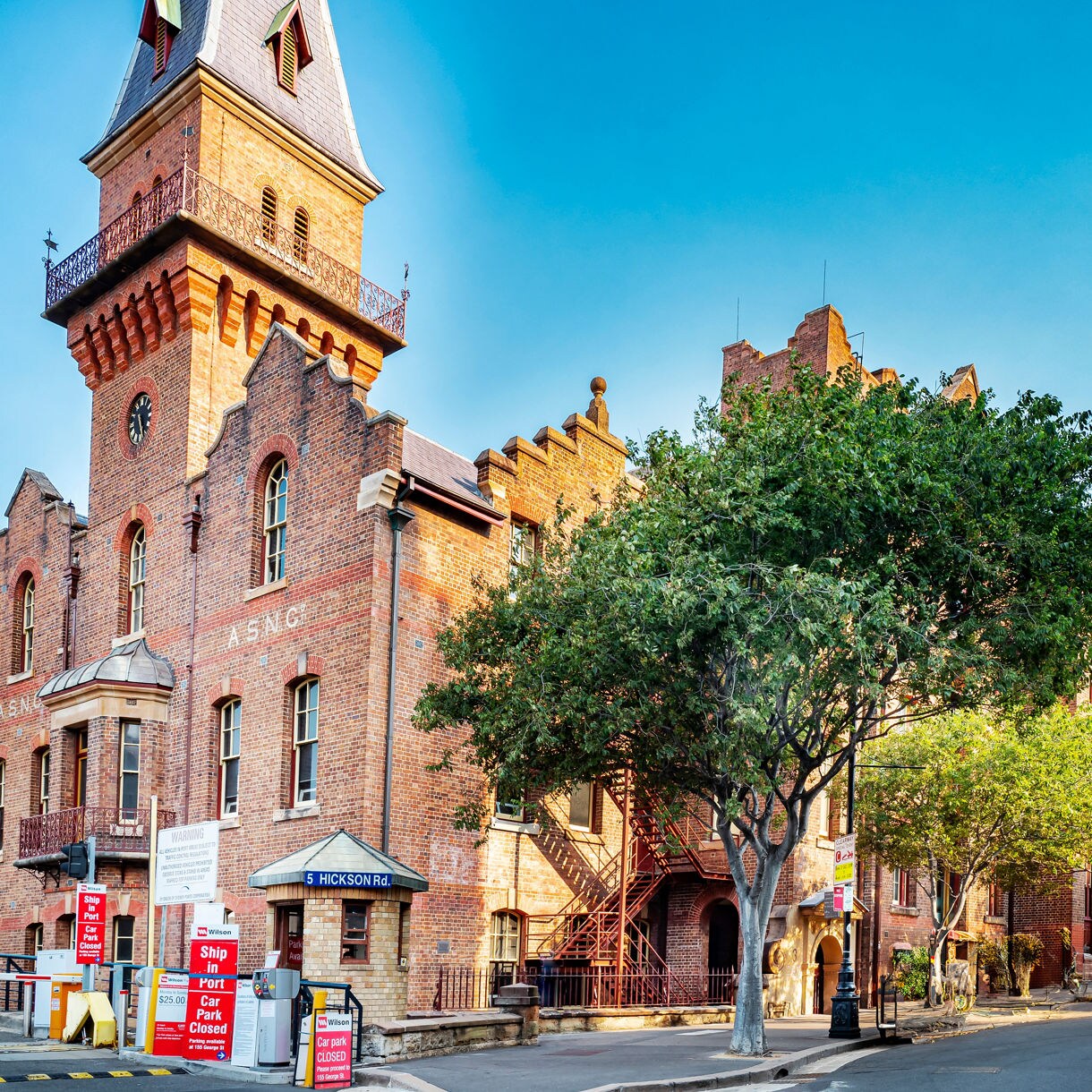 Street view of The Rocks district in Sydney with red-brick heritage buildings, a tall clock tower, leafy trees and quiet sidewalks under a clear blue morning sky.