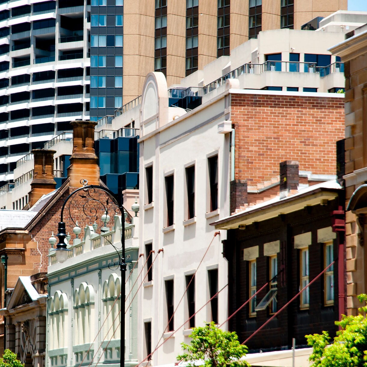 Close-up street view of The Rocks in Sydney featuring historic brick and stone facades in the foreground with tall contemporary skyscrapers rising directly behind them.