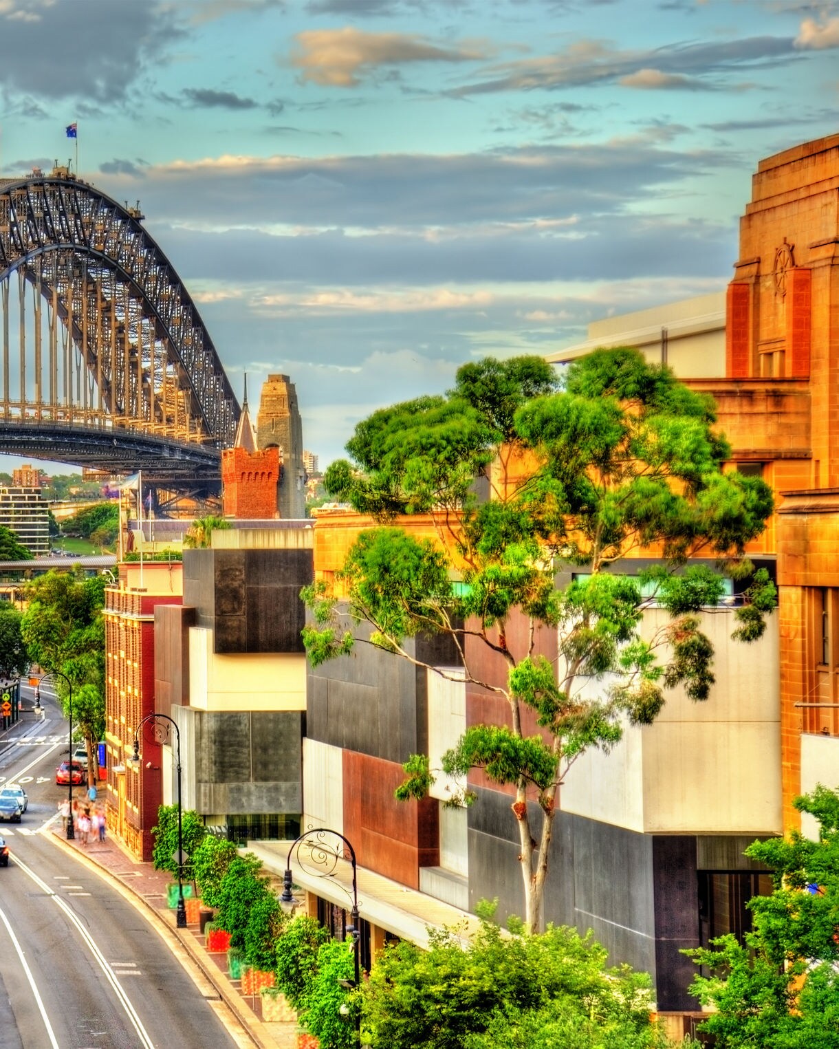 Vibrant view of The Rocks district in Sydney with colorful heritage buildings, leafy streets and the Sydney Harbour Bridge arching overhead under a partly cloudy sky.