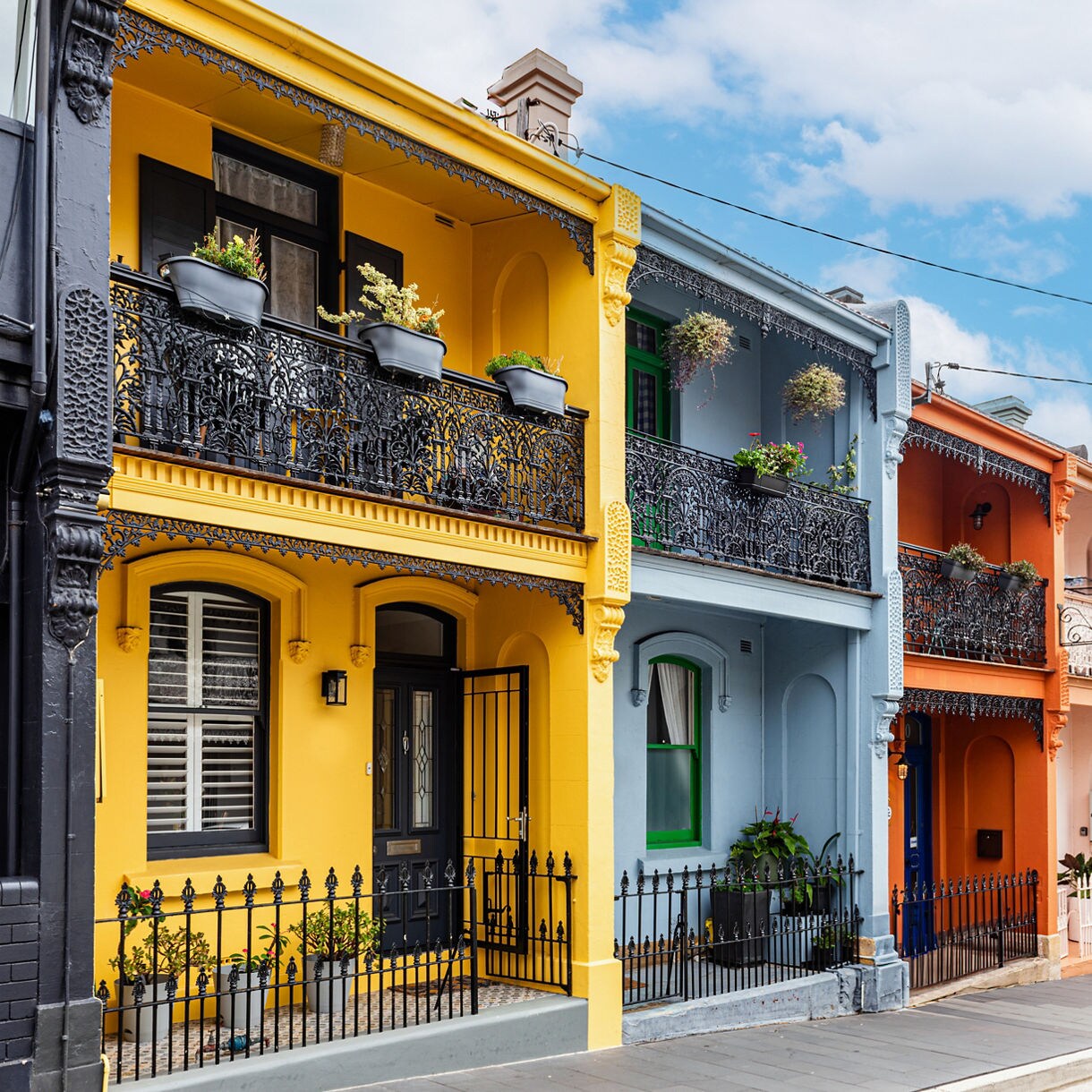 Row of vibrant Victorian terrace houses in Sydney painted yellow, blue, orange and soft pastels, each with decorative wrought-iron balconies, potted plants and a clean sidewalk under a partly cloudy sky.