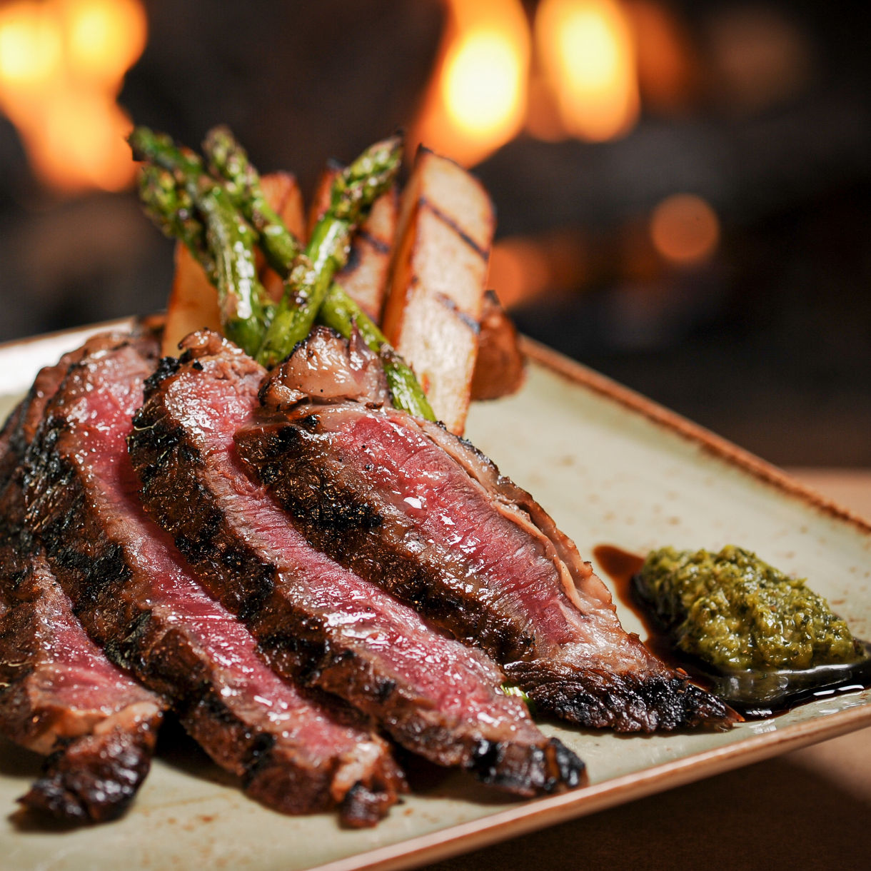 A sliced medium-rare steak fanned on a white plate with a dark seared crust, accompanied by grilled asparagus spears and roasted potatoes, with warm candlelight glowing softly in the blurred background.
