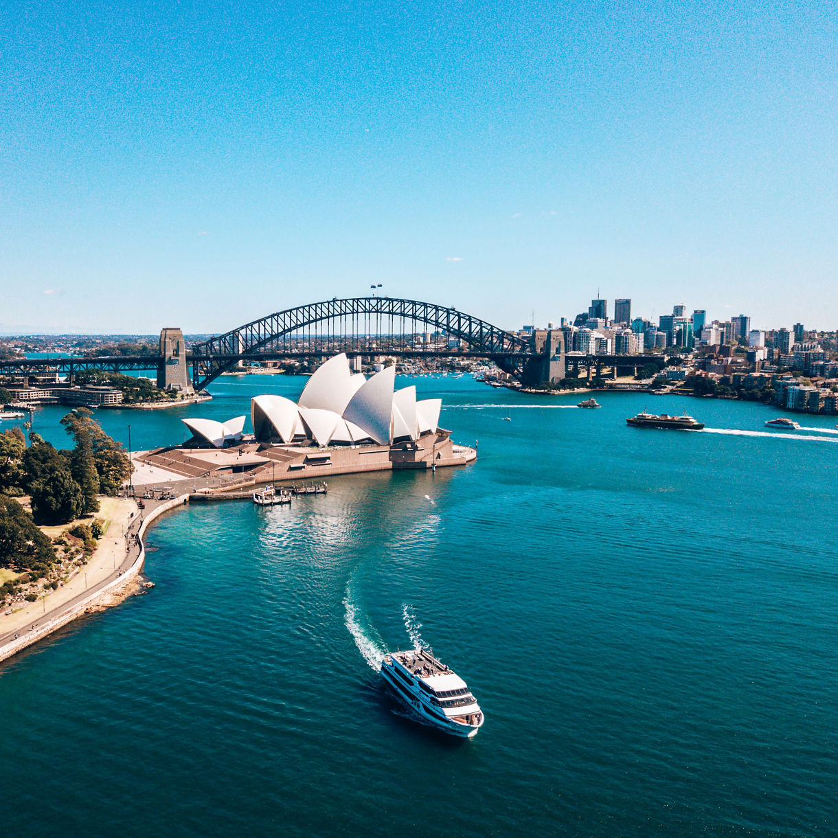 Aerial view of Sydney Harbour with a boat cruising past the Opera House and Harbour Bridge, framed by parkland and the city skyline.