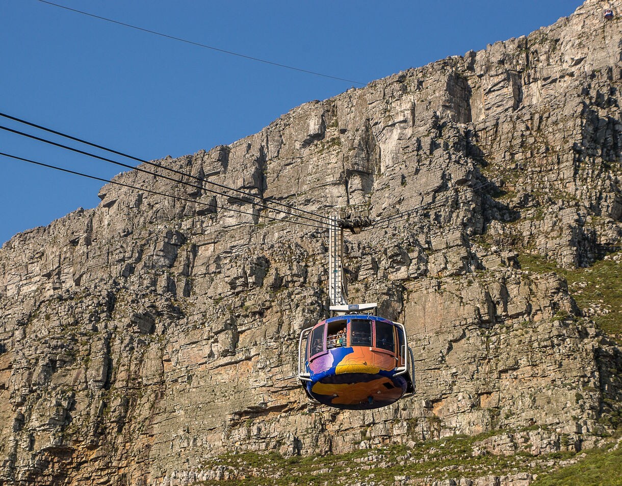 A brightly painted cable car ascending in front of a steep rocky mountain face.