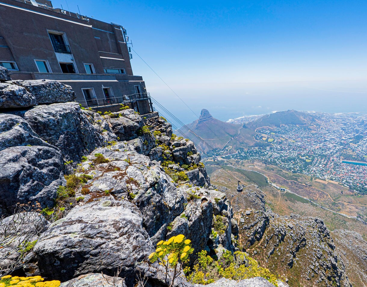 View from a rocky cliff on Table Mountain overlooking Cape Town, with a cable car descending and Lion’s Head visible in the distance.